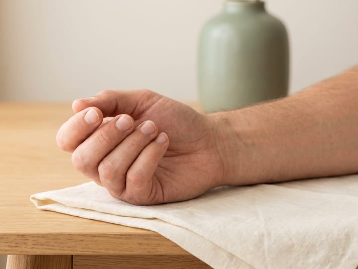 Mão adulta em repouso sobre um tecido de linho claro, em uma mesa de madeira, com luz natural suave.